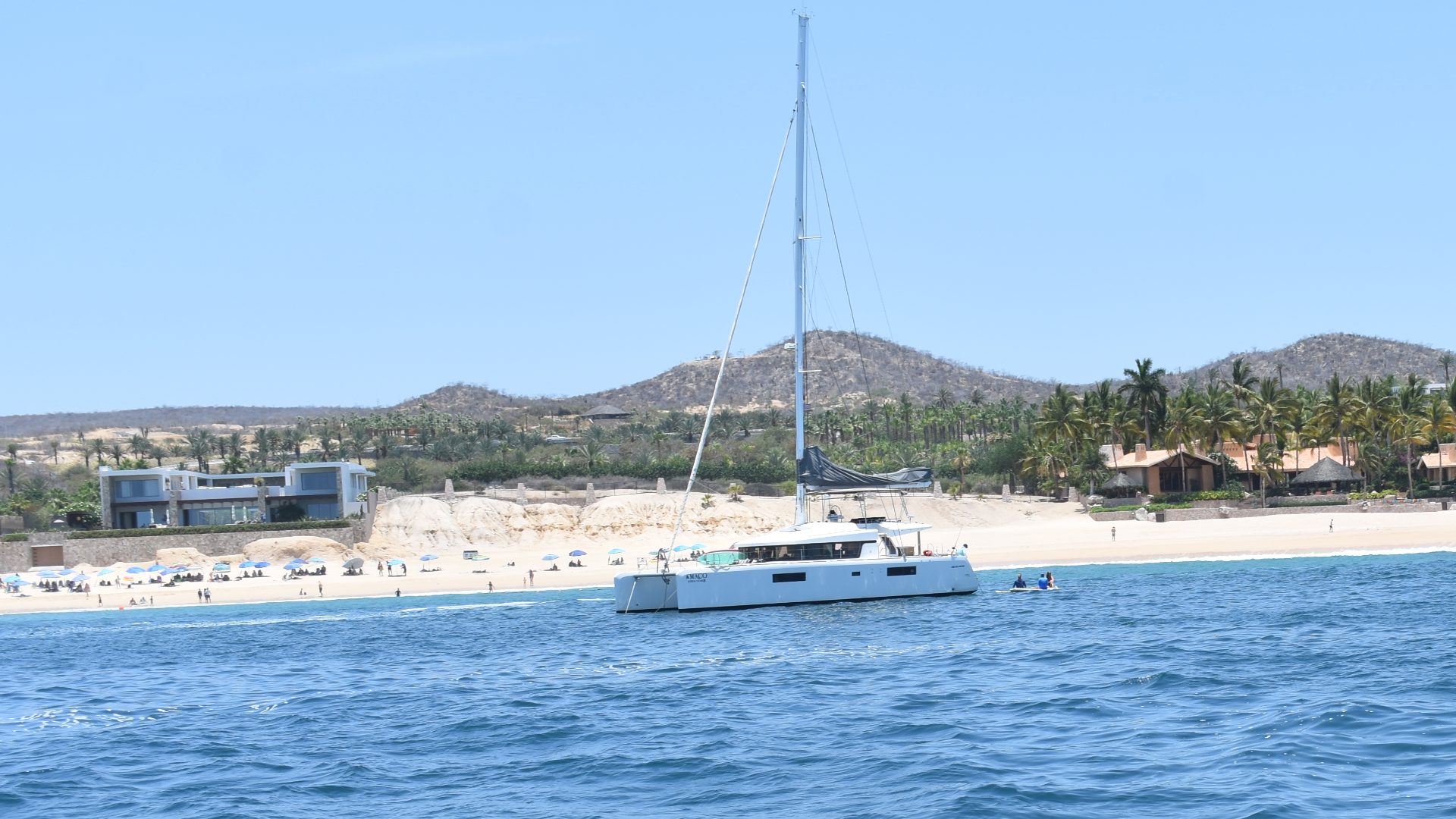 Sailing catamaran at anchor off a sandy beach on the Los Cabos coast, with desert hills behind