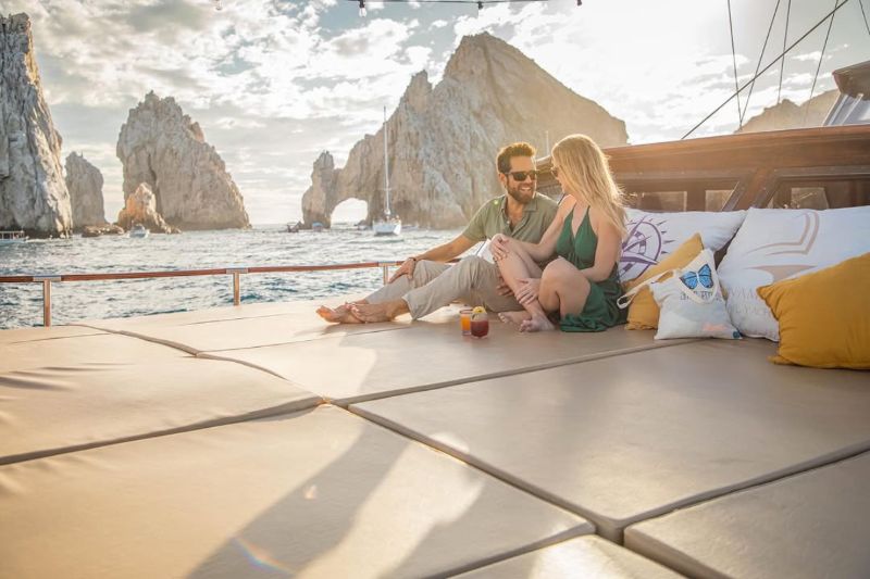 Couple relaxing on the cushioned deck of a sailing vessel with El Arco behind them