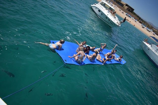 Guests relaxing on a floating mat in a calm bay beside a yacht