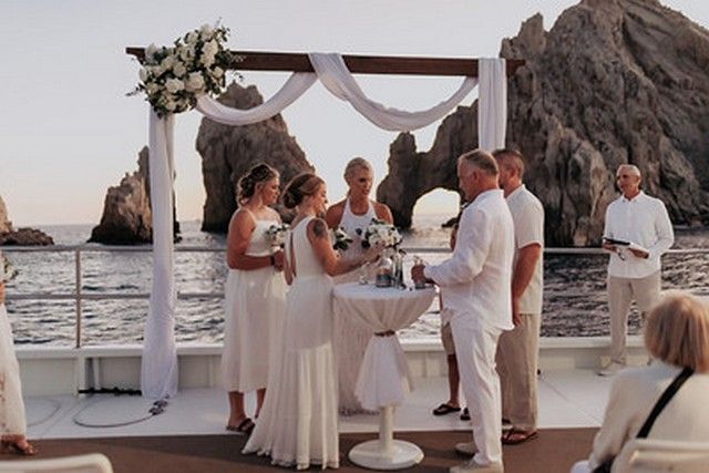 Couple exchanging vows under a floral arch on a catamaran with El Arco behind them