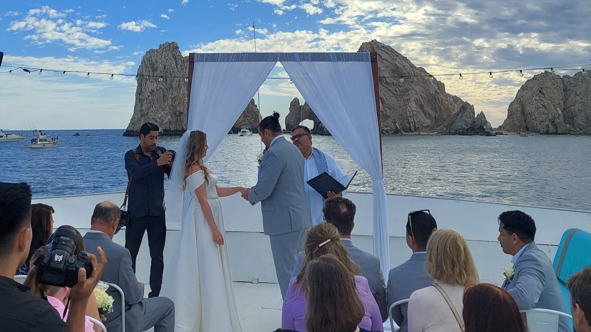 Bride and groom exchanging vows under a white arch on a catamaran deck, with Land's End behind them